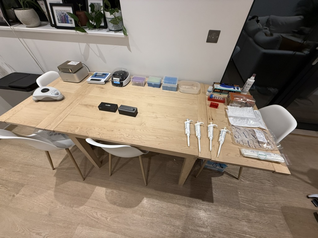 A dining-room table laid out with the full home-sequencing kit: pipettes, heat block, centrifuge, vortex, MinION boxes, tip boxes, reagent packaging.