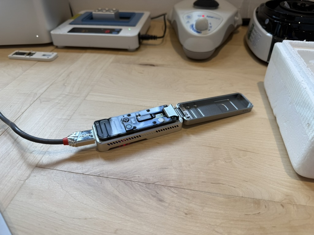 The MinION with its lid hinged open, flow cell seated, light shield removed, sitting on a kitchen table alongside a heat block, vortex, and centrifuge.